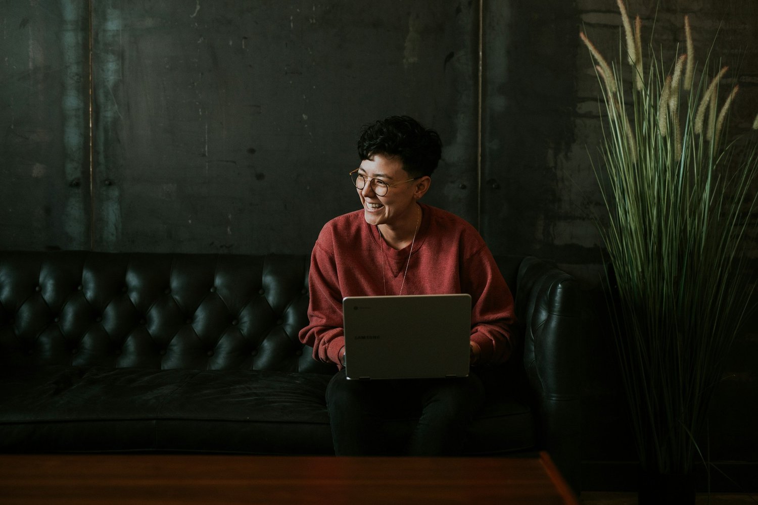 Person smiling at a laptop on a leather sofa in a dark lounge
