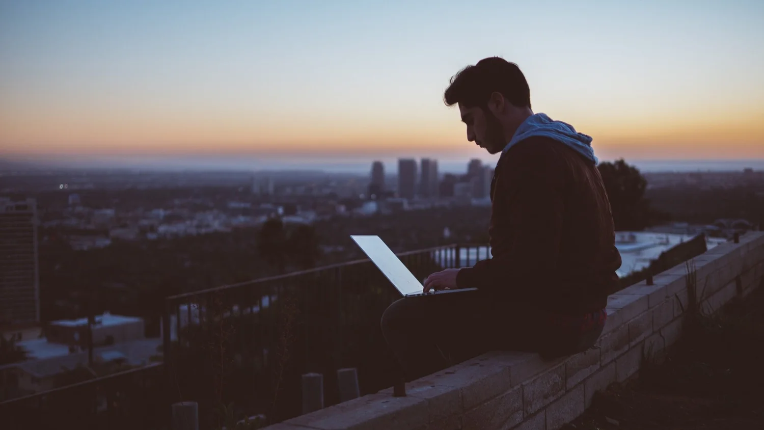 Remote worker on a laptop at a rooftop at dusk with a city skyline behind